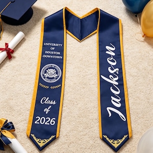 May include: A navy blue graduation stole with gold trim. The left side features the text University of Houston-Downtown, a circular university seal, and the text Class of 2026. The right side features the name Jackson in large white script.