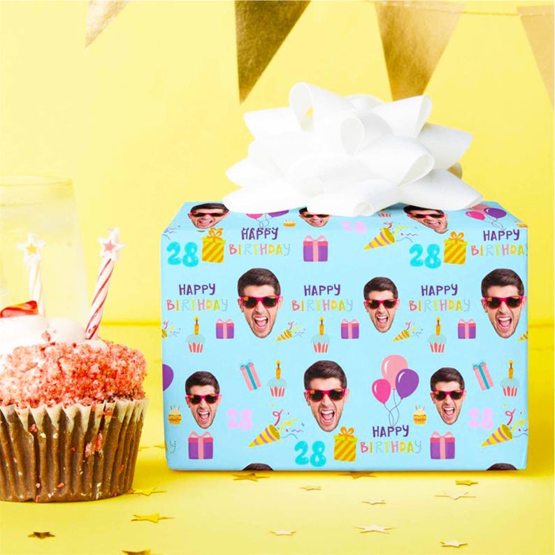 May include: A wrapped gift with a white bow on top, featuring a light blue wrapping paper with a repeating pattern of faces, gifts, and the text "Happy Birthday" and the number "28". A cupcake with candles is in the foreground.