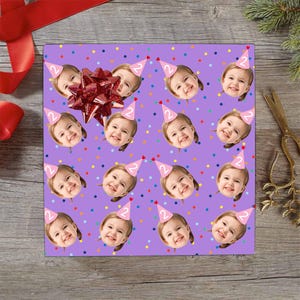 May include: Purple wrapping paper with a repeating pattern of a smiling child wearing a pink party hat with the number 2. The paper is decorated with colorful polka dots and a red bow.