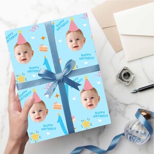 May include: A wrapped gift with a light blue background featuring a repeating pattern of a baby's face wearing a pink party hat, candles, cake slices, and the words "Happy Birthday." The gift is tied with a blue ribbon.