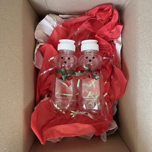 May include: Two clear plastic bear-shaped bottles with white caps, decorated with red and green ribbons, in a cardboard box with red tissue paper and silver ribbon. The bottles are empty.