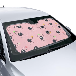 Könnte beinhalten: Ein rosa Autosonnenblende mit einem sich wiederholenden Muster aus schwarzen Cartoon-Charakteren, die bunte Kugeln halten. Die Sonnenblende ist in einem Auto installiert und bietet Schatten vor der Sonne.