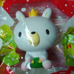 May include: White plush toy bear wearing a gold crown and green pants. The bear is sitting on a red and white background with green and white Christmas trees. The text "Squishy Good" is visible in the top right corner of the image.