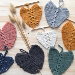 Könnte beinhalten: Eine Sammlung handgefertigter Makramee-Blattdekorationen in verschiedenen Farben, darunter Rosa, Senfgelb, Grau, Weiß, Marineblau, Dunkelgrau und Türkis. Jedes Blatt ist an einem kleinen Holzstab zum Aufhängen befestigt. Die Blätter sind auf einer hellen Holzoberfläche angeordnet.