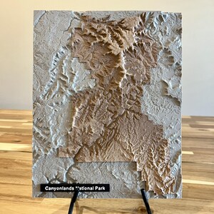 May include: A 3D topographic map of Canyonlands National Park, featuring detailed relief patterns in shades of beige and gray. The map is mounted on a rectangular base and displayed on a black metal stand. The text "Canyonlands National Park" is visible.