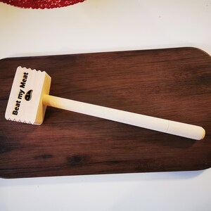 May include: A wooden meat mallet with a white handle. The mallet has a textured surface and the words "Beat my Meat" are printed on the top.