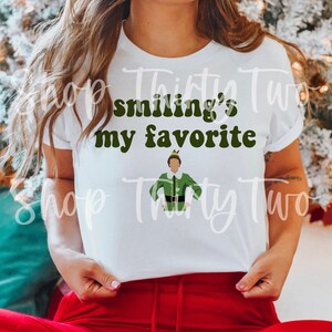Op de afbeelding: Wit T-shirt met de tekst "Smiling's my favorite" in groene letters met een cartoon afbeelding van Buddy de Elf uit de film Elf.