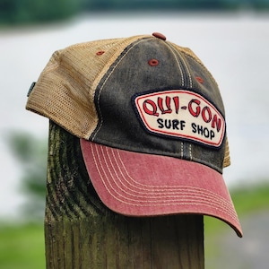 May include: A baseball cap featuring a dark charcoal front panel, a red brim, and tan mesh back panels. A patch on the front displays the text "QUI-GON SURF SHOP" in red and black lettering. The cap has a button on top and visible stitching on the brim.