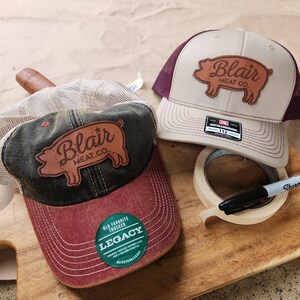 May include: Two brown and beige trucker hats with a brown leather patch featuring a pig and the text "Blair Meat Co.". The hats are on a wooden surface with a roll of tape and a black marker.
