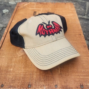 May include: A beige and black trucker hat with a black bat graphic and the text "Mothman" in red letters.