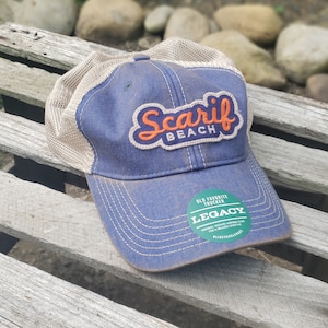 May include: Blue trucker hat with "Scarif Beach" patch in orange and white. The hat has a mesh back and a curved brim. The hat also has a green sticker that says "Legacy".