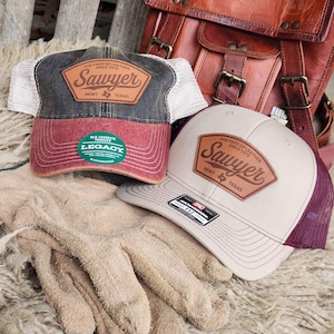 May include: Two brown and beige trucker hats with a brown leather patch that says "The Face of Leather Since 1973 Sawyer Newt Texas". The hat on the left has a red and black mesh back and a green sticker that says "Old Favorite Trucker Legacy". The hat on the right has a maroon and beige mesh back.