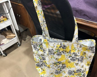 May include: A white tote bag with a yellow and black honeycomb pattern and floral design. The bag has black straps and is hanging on the back of a black office chair.