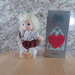 May include: A Precious Moments doll with blonde hair, wearing a white dress with red plaid accents. The doll is holding a heart-shaped sign that reads "Peace On Earth Begins In The Heart." A gray box with the words "Inspirations from the Heart" is next to the doll.