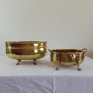 May include: Two antique brass planters. The larger planter has a rope-like trim and rests on three claw feet. The smaller planter has handles and decorative accents. Both planters have a hammered texture.