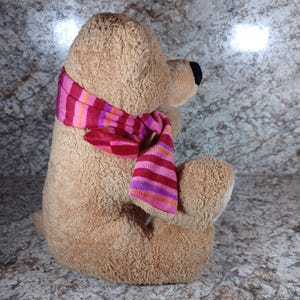 K&ouml;nnte beinhalten: Ein sitzender Teddyb&auml;r mit hellbraunem Fell und schwarzer Nase. Der B&auml;r tr&auml;gt einen gestreiften Schal in Rosa, Rot und Orange. Das Kuscheltier ist weich und f&uuml;r Kinder geeignet.