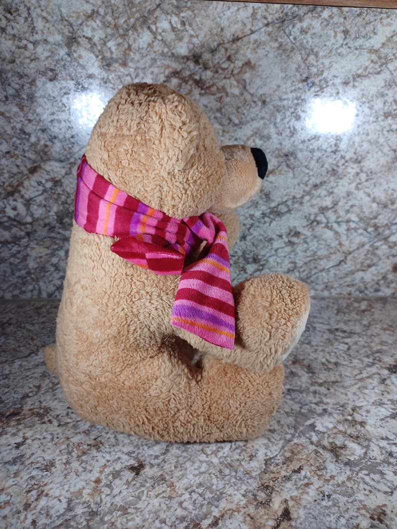 K&ouml;nnte beinhalten: Ein sitzender Teddyb&auml;r mit hellbraunem Fell und schwarzer Nase. Der B&auml;r tr&auml;gt einen gestreiften Schal in Rosa, Rot und Orange. Das Kuscheltier ist weich und f&uuml;r Kinder geeignet.