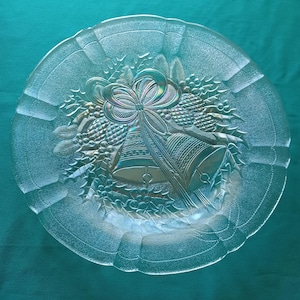 May include: Clear glass plate with embossed Christmas bells, bow, and holly design. The plate has a scalloped edge and is set against a teal background. A festive decorative piece for serving holiday treats.