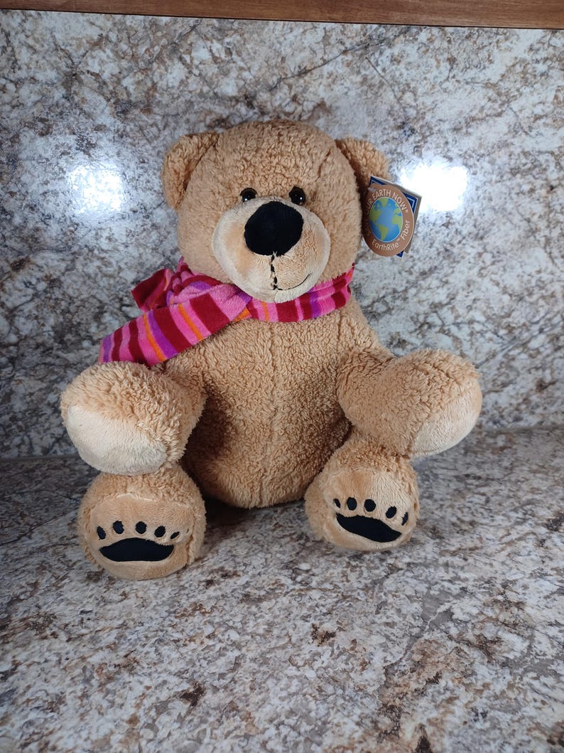 K&ouml;nnte beinhalten: Ein hellbrauner Teddyb&auml;r mit schwarzer Nase und Augen sowie einem rosa-rot gestreiften Schal. Der B&auml;r hat schwarze Tatzen und ein Etikett mit der Aufschrift "Earth Now".