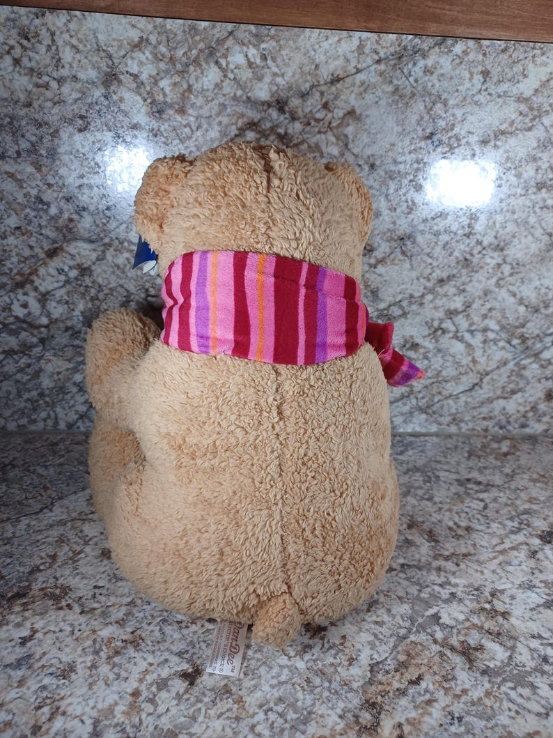 K&ouml;nnte beinhalten: Ein hellbrauner Teddyb&auml;r mit einem gestreiften Schal in Rosa, Rot und Lila. Das Stofftier ist von hinten zu sehen, mit dem Schal um den Hals. Das Fell des B&auml;ren ist strukturiert.