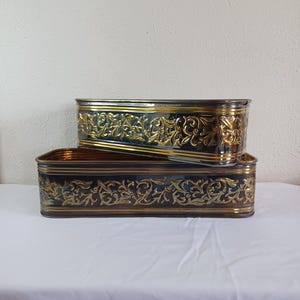 May include: Two rectangular metal planters stacked on top of each other. The planters have a dark blue base with gold-colored floral scrollwork and trim. The planters are sitting on a white surface.
