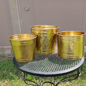May include: Three gold-colored metal planters of varying sizes. The smallest has vertical ridges, the middle one has a diamond pattern, and the largest has a hammered texture. They sit on a black metal table with a diamond-shaped mesh top, with grass in the background.