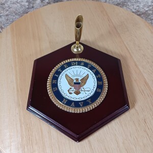 May include: A hexagonal dark wood desk pen holder featuring a centered circular emblem of the United States Navy. The emblem displays a golden eagle, a shield, and anchors surrounded by a rope-textured gold border. A gold metal pen socket is attached.
