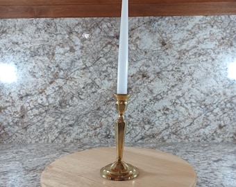 Puede incluir: Un candelabro de latón con una vela cónica blanca. El candelabro tiene una base detallada y un diseño esbelto. La vela está centrada en una tabla de madera redonda.