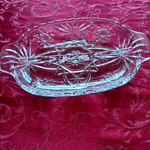 May include: Clear glass serving dish with starburst and geometric designs. The oval-shaped dish has handles on each end and is set against a red background. The dish is likely used for serving food.