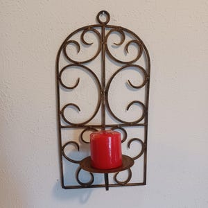 May include: A wall-mounted candle holder featuring a dark bronze-toned metal frame with an arched top and decorative scrollwork patterns. A circular platform extends from the lower section, supporting a single cylindrical red wax candle.