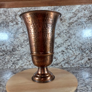 May include: A hammered copper vase with a flared top and a pedestal base. The vase has a warm, reddish-brown color and a textured surface. It is displayed on a round wooden surface.