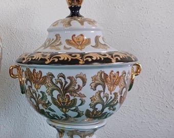 May include: A white and gold decorative lidded bowl with a pedestal base. The bowl features ornate gold floral designs and a black band around the rim. The lid is topped with a gold finial. The bowl has gold handles.