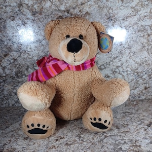 May include: A plush teddy bear with a light brown fur, black eyes and nose, and a pink and red striped scarf. The bear has paw prints on its feet and a tag that reads "Earth Now".