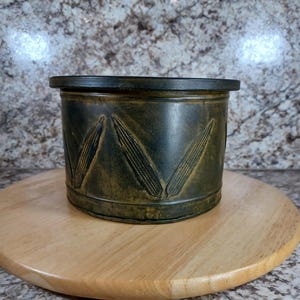 May include: A cylindrical, dark green metal container with embossed corn designs. The container has a dark rim and sits on a round, light-colored wooden surface. The container's exterior has a distressed, aged appearance.