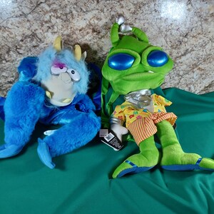 May include: Two plush toys: a blue monster with yellow horns and a green alien with large blue eyes. The monster has a furry body and a cartoonish face. The alien wears a yellow shirt and orange striped shorts. Both are on a green surface.