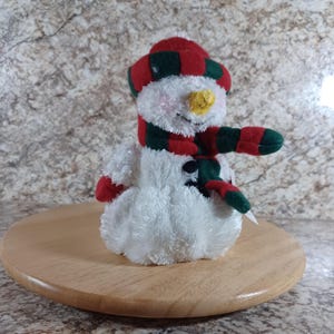 May include: A plush snowman figurine with a red and green plaid hat and scarf. The snowman has a yellow nose, black button eyes, and red mittens. It is sitting on a round wooden base.
