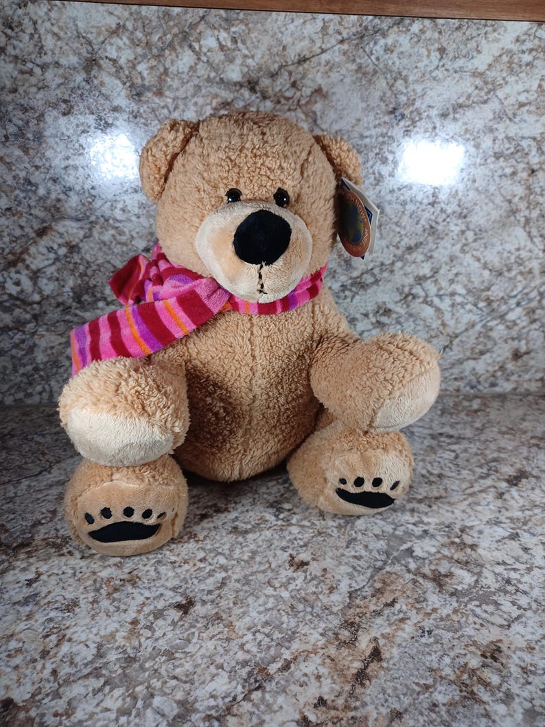 K&ouml;nnte beinhalten: Ein brauner Teddyb&auml;r mit schwarzer Nase und Augen, der einen rosa-rot gestreiften Schal tr&auml;gt. Der B&auml;r hat schwarze Pfotenabdr&uuml;cke auf seinen F&uuml;&szlig;en und ein Etikett an seinem Ohr. Ein kuscheliges Stofftier.