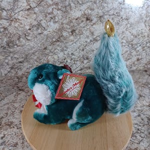 May include: A teal plush squirrel with a fluffy tail and a gold-colored acorn on top. The squirrel has a red bow and a tag that reads "Winter Magic". The plush toy is sitting on a wooden round base.
