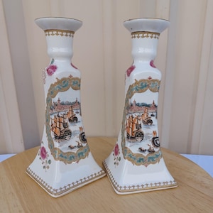 May include: A pair of white porcelain candle holders with gold trim and a scene of ships sailing in a harbor. The candle holders are square shaped with a decorative design.