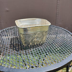 May include: A metallic gold-toned planter with a square shape and rounded corners. The exterior features a textured, woven-style pattern on the lower section and horizontal ribbed detailing around the upper rim, creating a reflective surface.