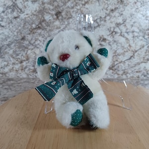 May include: A small, white teddy bear with a red nose, green accents on the ears and paws, and a dark green bow with a white pattern. The bear is made of soft, plush material.