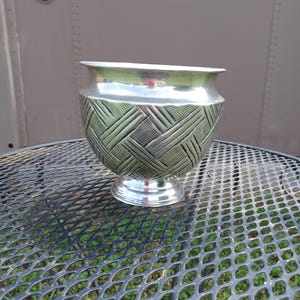 May include: A silver-colored metal bowl with a textured, geometric pattern on the lower half. The bowl has a wide, open top and sits on a small, round base. The bowl is displayed on a black metal mesh table.