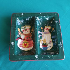 May include: A rectangular, divided ceramic serving dish with a green background and a red border. The dish features two snowman chefs with cooking utensils and snowflake accents, perfect for holiday entertaining.
