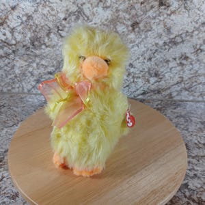 May include: A plush yellow chick with orange feet and a matching beak. The chick has a fluffy appearance and wears a sheer pink and yellow bow. A red tag is attached to the chick's side.
