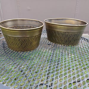 May include: Two bronze-colored metal planters with a woven basket design. The planters have a rounded oval shape and are sitting on a metal mesh table. The planters are suitable for outdoor or indoor use.