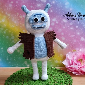 May include: A crocheted blue and white monster toy wearing a brown waistcoat. The toy is standing on a green crocheted doily. The background is a rainbow ombre with white sparkles. The text "Adri's Designs *crafted gifts*" is in the top right corner of the image.