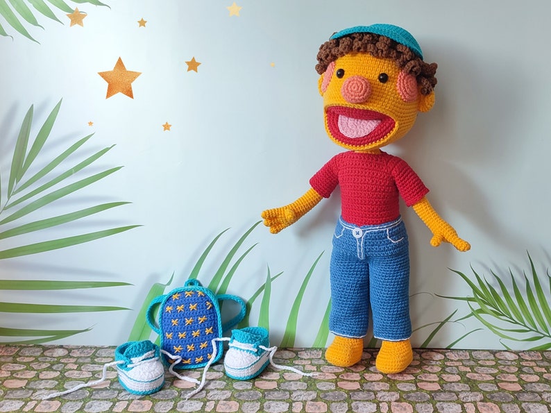 Puede incluir: Una mu&ntilde;eca de crochet que lleva una camisa roja, pantalones vaqueros azules y un sombrero azul. La mu&ntilde;eca lleva una mochila azul con estrellas amarillas. La mu&ntilde;eca est&aacute; de pie sobre un suelo de piedra con plantas verdes en el fondo.