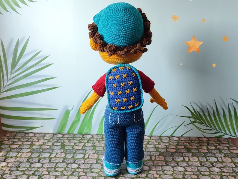Puede incluir: Una mu&ntilde;eca de crochet con un sombrero azul, una camisa roja, pantalones vaqueros azules y una mochila azul con estrellas amarillas. La mu&ntilde;eca est&aacute; de pie en un camino de piedra.