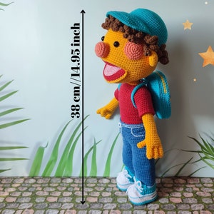 Puede incluir: Un juguete de peluche de crochet de un ni&ntilde;o que lleva un sombrero azul, una camisa roja, pantalones vaqueros azules y una mochila azul. El juguete mide 38 cm de alto.