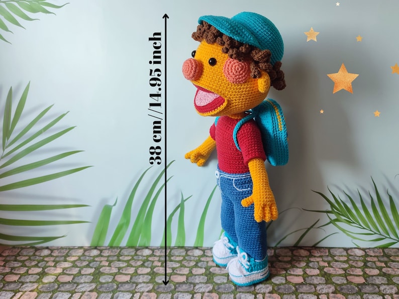 Puede incluir: Un juguete de peluche de crochet de un ni&ntilde;o que lleva un sombrero azul, una camisa roja, pantalones vaqueros azules y una mochila azul. El juguete mide 38 cm de alto.